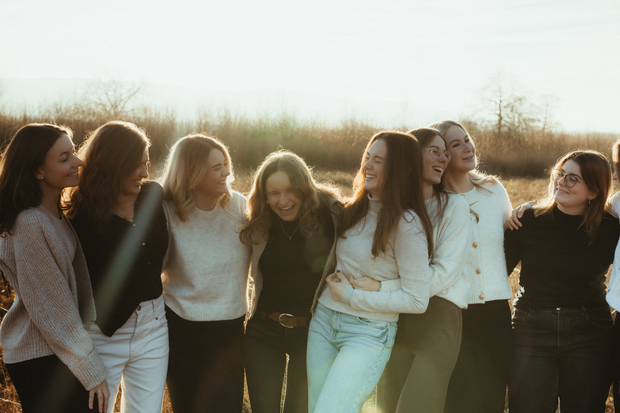 Junge Frauen lachen gemeinsam in der Abendsonne und umarmen sich auf einer Wiese.