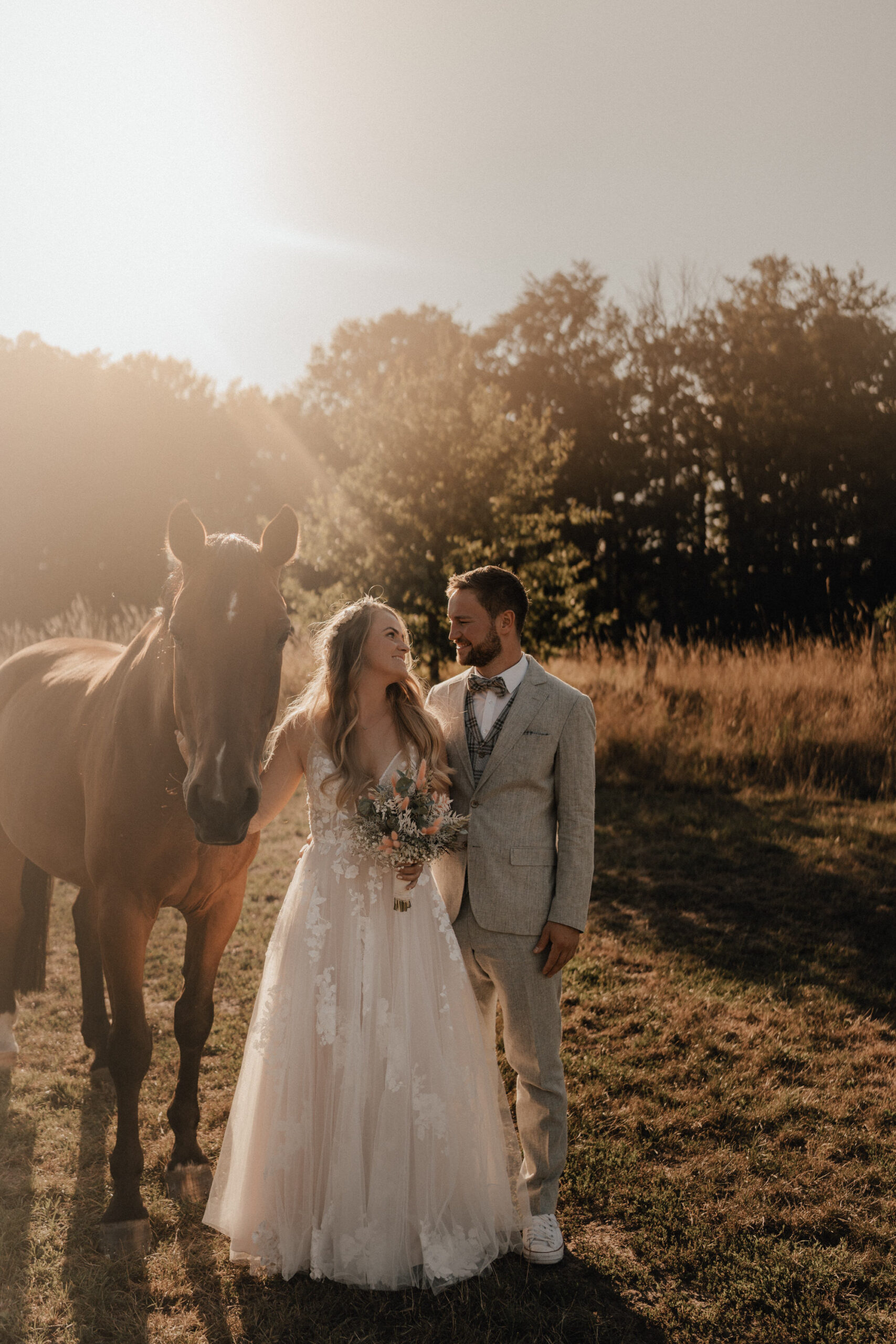 Brautpaar in Boho-Outfits steht neben einem Pferd auf einer Wiese im Sonnenuntergang – emotionale Hochzeitsfotografie von Laura Viermann.