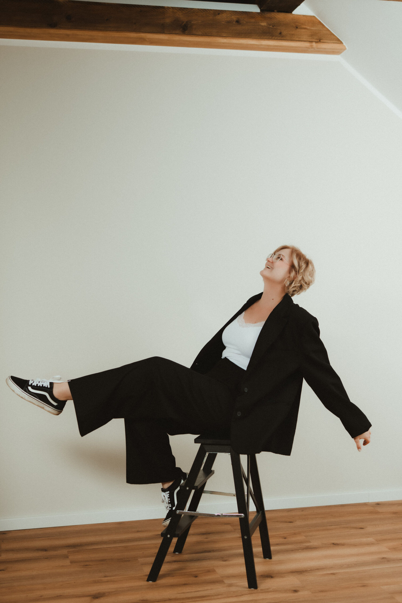 Laura Viermann sitzt lässig auf einem Hocker, trägt ein schwarzes Outfit mit Sneakers und lacht – ein modernes, unkonventionelles Portrait.