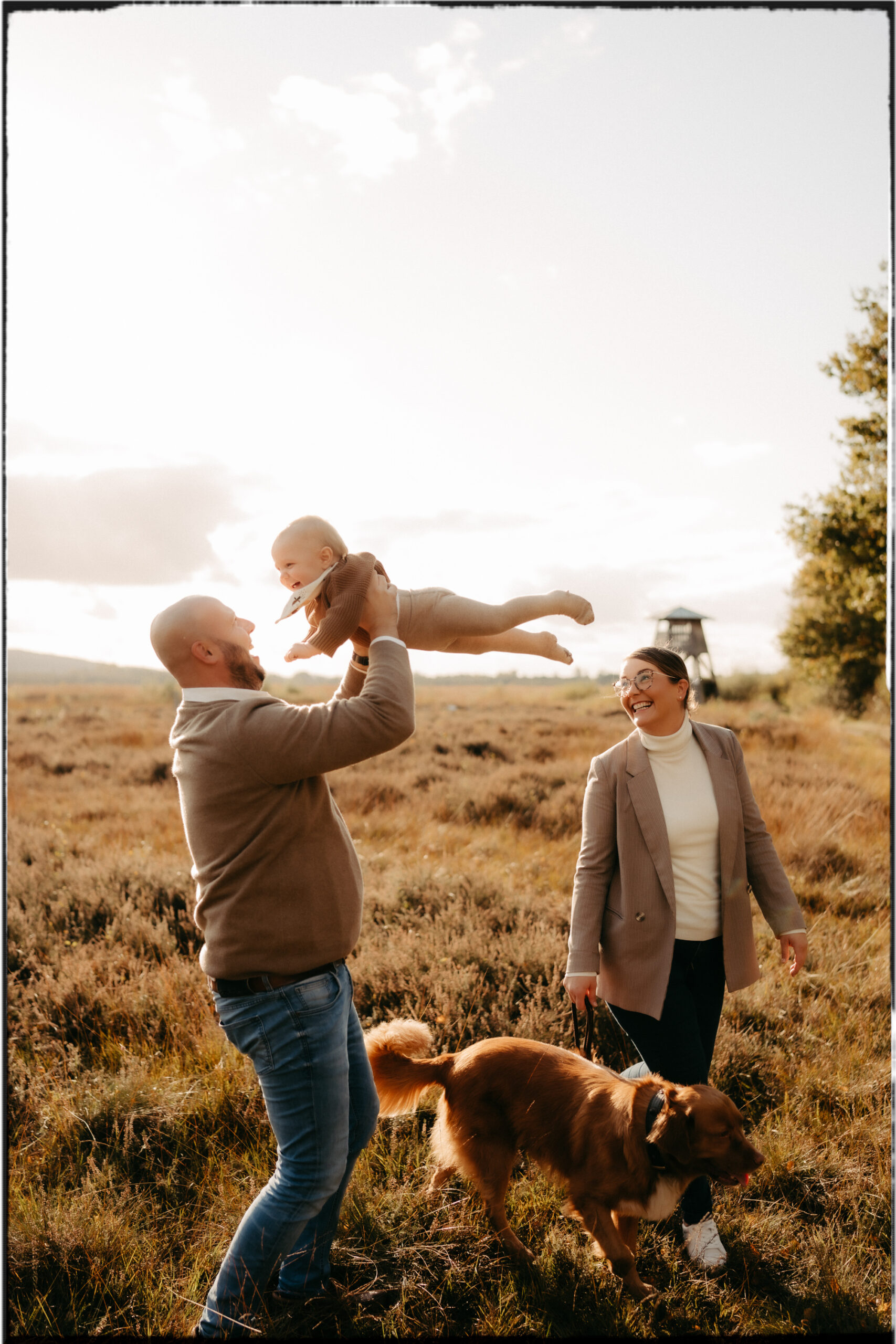 Ein Vater wirft sein Baby lachend in die Luft, die Mutter schaut zu und hält den Familienhund an der Leine – Outdoor-Familienfoto mit Sonnenlicht.